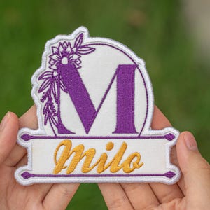 May include: Embroidered patch featuring the letter "M" in purple, with a floral design. The name "Milo" is embroidered in gold script below the letter. The patch has a white background and a decorative border.