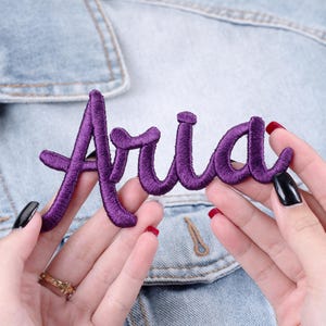 May include: A purple embroidered patch with the name "Aria" in a cursive script. The patch is held against a light blue denim jacket, showcasing the vibrant color and design of the patch.