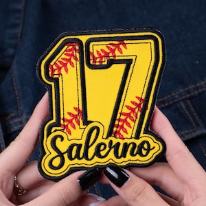 May include: A yellow and black embroidered patch with the number 17 and the name "Salerno". The number is outlined in black and filled with yellow fabric. Red stitching resembling baseball seams is visible within the numbers. The patch is held against a denim background.