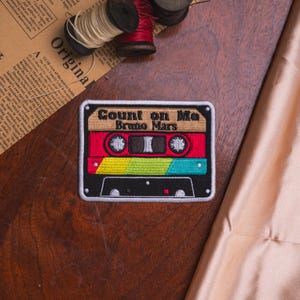 May include: A rectangular embroidered patch shaped like a cassette tape with a white border. The design features a tan top section with the black text Count on Me Bruno Mars, a red middle section with two circular reels, and a black bottom section.