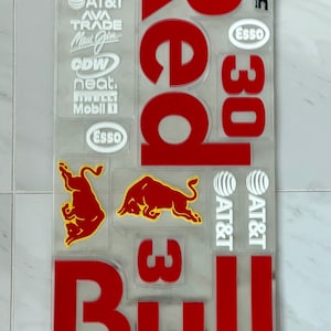 May include: A clear decal sheet featuring various red and white sponsor logos, including Red Bull text, two red bull silhouettes, the number 30, and multiple brand names like Visa, AT&T, and Esso. A 1cm x 1cm square reference guide is printed at the top.