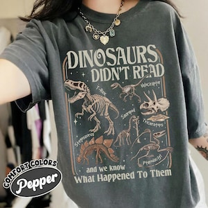 May include: Dark gray t-shirt with the text "DINOSAURS DIDN'T READ" and illustrations of dinosaur skeletons. The shirt also features the text "and we know What Happened To Them" and the Comfort Colors Pepper logo.