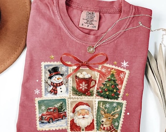 Puede incluir: Una camiseta de manga corta color carmesí con un estampado gráfico de cinco ilustraciones estilo sello postal que representan un muñeco de nieve, una taza de cacao, un árbol de Navidad, un camión rojo, Santa Claus y un reno, unidos por un lazo rojo.
