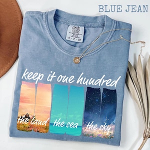 May include: Blue jean colored t-shirt with the text "keep it one hundred". The shirt features colorful graphics of the land, sea, and sky. A gold necklace with a pendant rests on the shirt.