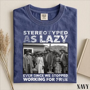 Puede incluir: Una camiseta azul marino de cuello redondo con una fotograf&iacute;a hist&oacute;rica en blanco y negro de un grupo de personas. El texto STEREOTYPED AS LAZY est&aacute; impreso sobre la imagen y EVER SINCE WE STOPPED WORKING FOR FREE debajo de la imagen.