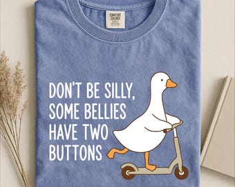 Puede incluir: Una camiseta color azul jean con un gráfico de un ganso blanco montado en un patinete. El texto dice: DON'T BE SILLY, SOME BELLIES HAVE TWO BUTTONS. La camiseta presenta una etiqueta de la marca Comfort Colors en el cuello y cuello redondo.