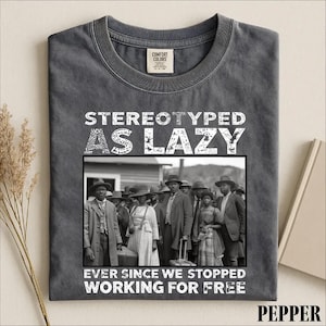 Puede incluir: Una camiseta gris carb&oacute;n con una fotograf&iacute;a hist&oacute;rica en blanco y negro de un grupo de personas con ropa formal. El texto "STEREOTYPED AS LAZY" aparece sobre la imagen y "EVER SINCE WE STOPPED WORKING FOR FREE" est&aacute; impreso debajo.