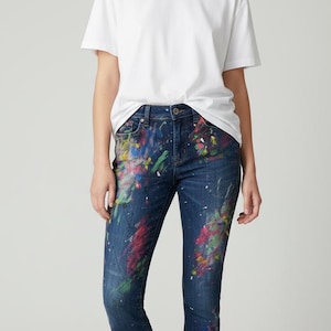 May include: A woman wearing a white t-shirt and dark blue jeans with colorful paint splatter designs. The jeans have a slim fit. The model is barefoot.