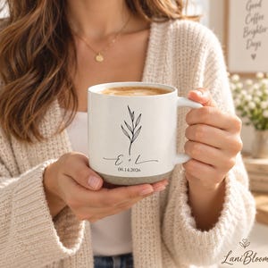 Könnte beinhalten: Ein weißer Keramikbecher mit gesprenkeltem grauen Boden, verziert mit einer schwarzen botanischen Strichzeichnung, den Buchstaben E + L und dem Datum 06.14.2026 auf der Vorderseite. Der Becher hat einen glatten Henkel und enthält ein hellbraunes Getränk.