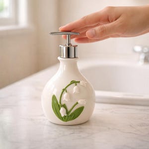 Puede incluir: Dispensador de jabón de cerámica blanca con bomba plateada. El dispensador presenta un diseño de lirios de los valles verdes y blancos. El dispensador está sobre una encimera de mármol.