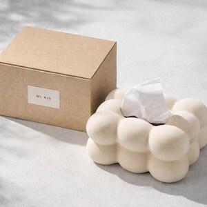 Puede incluir: Un dispensador de pa&ntilde;uelos de papel beige con un dise&ntilde;o redondeado &uacute;nico, junto a una caja de cart&oacute;n a juego. El soporte para pa&ntilde;uelos tiene una apariencia de burbujas, con un pa&ntilde;uelo blanco asomando. La caja de cart&oacute;n tiene el texto "MI KIS".