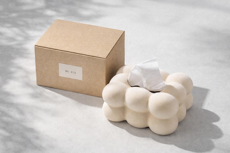 Puede incluir: Un dispensador de pa&ntilde;uelos de papel beige con un dise&ntilde;o redondeado &uacute;nico, junto a una caja de cart&oacute;n a juego. El soporte para pa&ntilde;uelos tiene una apariencia de burbujas, con un pa&ntilde;uelo blanco asomando. La caja de cart&oacute;n tiene el texto "MI KIS".