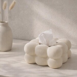Puede incluir: Una caja de pa&ntilde;uelos de color crema con forma de burbujas y pa&ntilde;uelos blancos. A la izquierda hay un jarr&oacute;n a juego con flores secas. Los objetos est&aacute;n sobre una superficie clara, creando una est&eacute;tica minimalista.