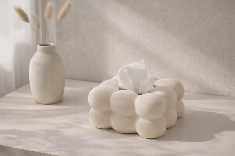 Puede incluir: Una caja de pa&ntilde;uelos de color crema con forma de burbujas y pa&ntilde;uelos blancos. A la izquierda hay un jarr&oacute;n a juego con flores secas. Los objetos est&aacute;n sobre una superficie clara, creando una est&eacute;tica minimalista.