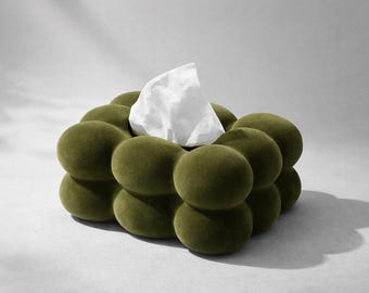 May include: A decorative tissue box cover in olive green, featuring a distinctive rounded shape. The cover has a textured, plush finish, with a white tissue visible. The design is modern and suitable for home decor.