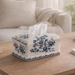 Puede incluir: Una caja para pañuelos rectangular con una base de color crema y un patrón floral azul detallado. El diseño incluye ilustraciones intrincadas de rosas y un borde decorativo en la parte inferior, con un pañuelo blanco sobresaliendo por arriba.