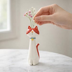 Peut inclure: Un vase en céramique blanc en forme de chat avec des détails rouges sur la tête et la queue. Le vase a un collier bleu avec un bouton jaune et contient de petites fleurs roses et blanches. Le vase est sur une surface blanche.
