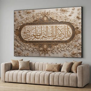 Può includere: Una grande opera d'arte incorniciata con calligrafia araba dorata su uno sfondo floreale beige. L'opera d'arte è esposta sopra un divano beige con diversi cuscini. L'estetica generale è elegante e decorativa.