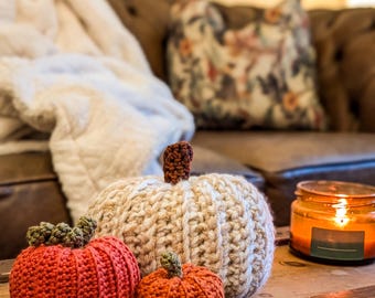 May include: Three handmade crochet pumpkins in varying sizes and colors sit on a wooden surface. The largest pumpkin is off-white, the medium one is orange, and the smallest is a darker orange. Each pumpkin has a crocheted stem.