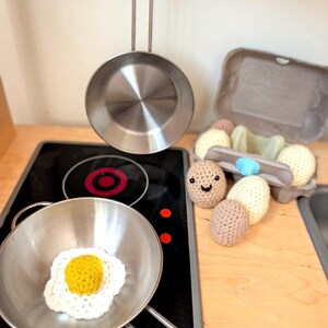 May include: A toy kitchen scene featuring a stainless steel frying pan with a crocheted fried egg. A second pan hangs above. Crochet eggs, some with smiling faces, sit near an egg carton on a wooden surface. A black stovetop is visible.