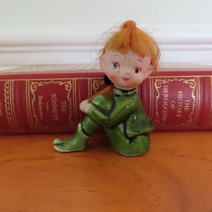 May include: A ceramic figurine of a seated elf with red hair and a green outfit. The elf is resting on a wooden surface in front of a red book with gold lettering. The book's title is "The Sonnets".