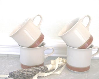 May include: Four white ceramic mugs with a brown stripe and base are stacked in pairs. Each mug has a curved handle. A bundle of dried lavender and a cream-colored ribbon are in the foreground.