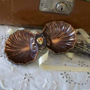 May include: A vintage copper-toned metal belt buckle with a seashell design. The buckle features an "I ❤️ NY" emblem in the center. The buckle rests on a white, embroidered doily with a brown suitcase in the background.