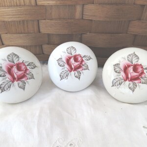 May include: Three round white ceramic cabinet knobs, each featuring a central illustration of a pink rose with grey-green leaves. The knobs have a smooth, glossy finish and a domed shape, displaying a consistent floral pattern across all three pieces.