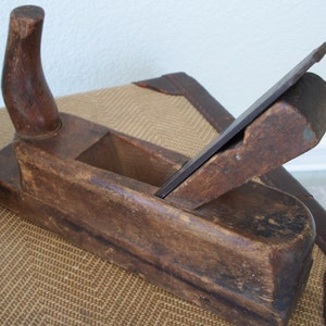 May include: A vintage wooden hand plane, showing signs of age and use. The plane has a dark brown finish with a curved handle and a metal blade. The blade is partially extended, and the body of the plane shows wear.