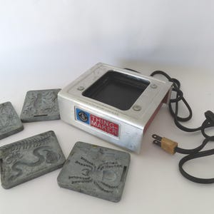 May include: A vintage Thingmaker toy heating unit with a silver metal exterior, a black central heating plate, and a black power cord. Included are four square grey metal molds featuring embossed designs of a spider, a snake, and other insects.