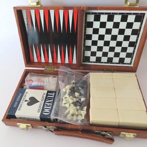 May include: A brown faux leather game case containing a backgammon board and a chessboard on the interior lid. The base holds a deck of Tuxedo playing cards, a plastic bag of small chess pieces, dice, and a set of cream-colored dominoes.