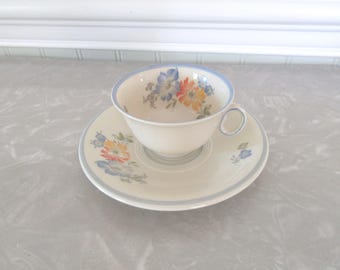 May include: A cream-colored ceramic teacup and saucer set featuring a delicate floral pattern with blue, orange, and yellow flowers. The rim of both the cup and the saucer is accented with a thin, light blue band, and the cup has a small handle.