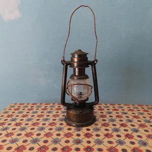 May include: A vintage-style metal lantern with a dark bronze finish, featuring a clear glass globe, a wire handle, and a central burner mechanism. The lantern has a sturdy circular base and a protective cage structure surrounding the glass globe.
