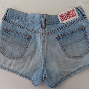 May include: Light blue denim shorts featuring a classic five-pocket design with copper-colored stitching. A white rectangular patch with the red text BUNGO is sewn onto the back waistband. The denim has a faded wash and a standard button fly.