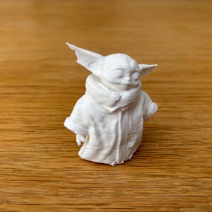 May include: A white figurine of a character with large ears and a hooded coat. The figure is sculpted with fine details, showcasing the texture of the clothing and the character's facial features. The figurine is set against a wooden background.