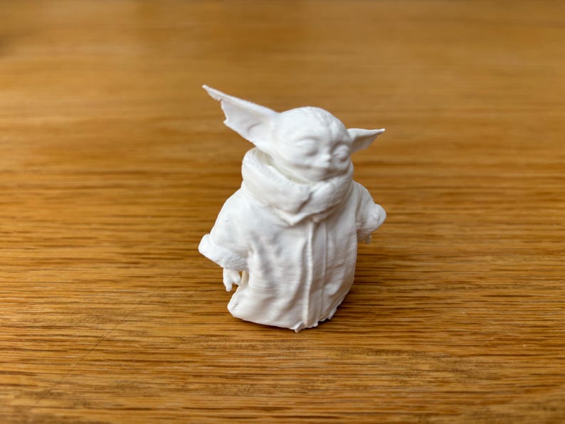 May include: A white figurine of a character with large ears and a hooded coat. The figure is sculpted with fine details, showcasing the texture of the clothing and the character's facial features. The figurine is set against a wooden background.