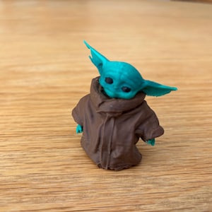 May include: A small figurine of a character with large ears and a brown coat. The figure has a teal-coloured head and ears, with a brown coat. The figurine is on a wooden surface.