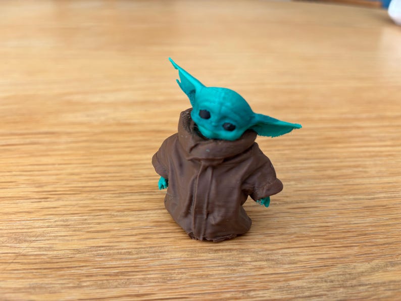 May include: A small figurine of a character with large ears and a brown coat. The figure has a teal-coloured head and ears, with a brown coat. The figurine is on a wooden surface.