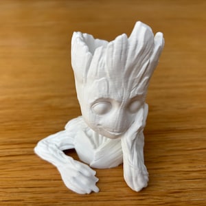 Könnte beinhalten: Ein weißer, 3D-gedruckter Pflanztopf in Form der Figur Groot aus Guardians of the Galaxy. Der Pflanztopf hat ein detailliertes Gesicht, Arme und ein strukturiertes, baumartiges Aussehen. Der Pflanztopf steht auf einer Holzoberfläche.