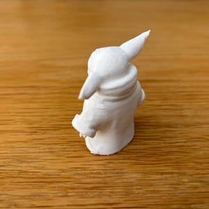 May include: A small, white figurine of a character with large ears and a hooded cloak. The figurine is standing on a wooden surface. The figure has a detailed texture and is likely a collectible item.