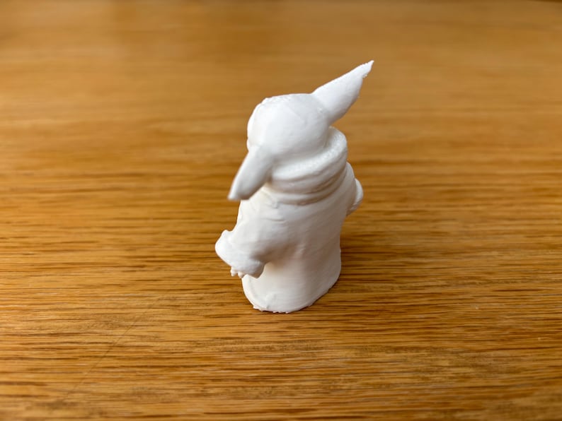 May include: A small, white figurine of a character with large ears and a hooded cloak. The figurine is standing on a wooden surface. The figure has a detailed texture and is likely a collectible item.