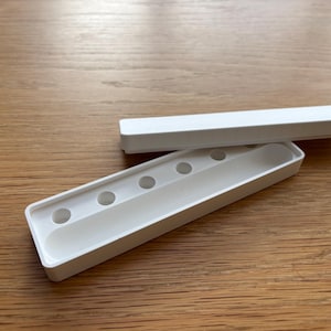 May include: A rectangular white plastic storage case with a removable lid. The base features a long, narrow recessed channel and five small circular indentations arranged in a single row, designed for holding small items or writing instruments.