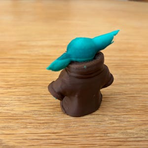 May include: A small, handmade figurine of a character with large ears and a brown coat. The figure has a teal-coloured head and is set against a wooden background. The figurine is a popular character from a science fiction franchise.