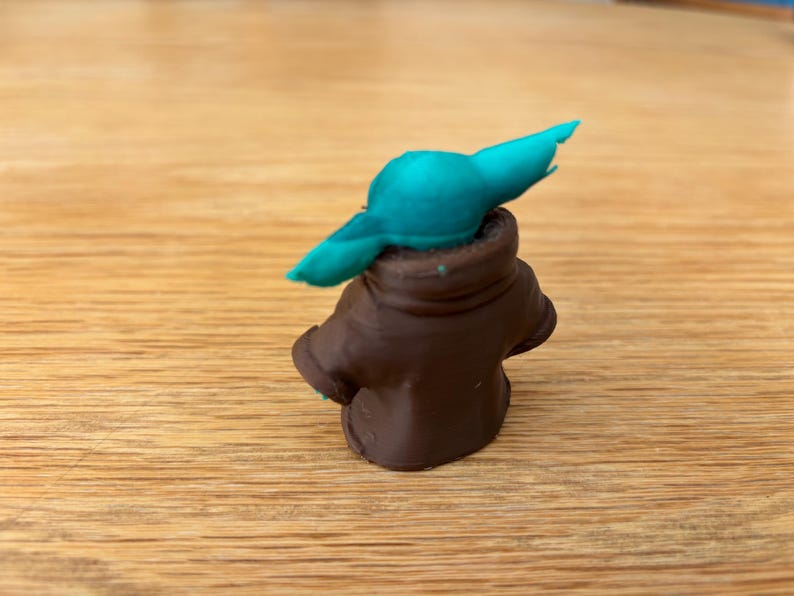 May include: A small, handmade figurine of a character with large ears and a brown coat. The figure has a teal-coloured head and is set against a wooden background. The figurine is a popular character from a science fiction franchise.