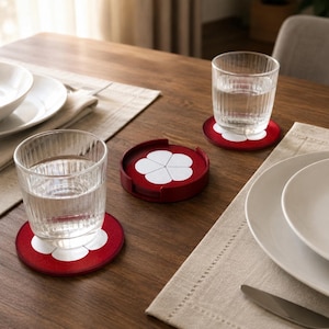 Pode incluir: Um conjunto de bases para copos redondas vermelhas com um design de flor branca de cinco pétalas. O conjunto inclui duas bases sob copos de água canelados e uma base guardada num suporte circular vermelho a condizer sobre uma mesa.