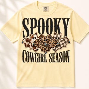 May include: A pale yellow short-sleeve t-shirt featuring the black text SPOOKY COWGIRL SEASON. The design includes three bat illustrations with cow print, leopard print, and checkered patterns, accented with small turquoise-colored studs.