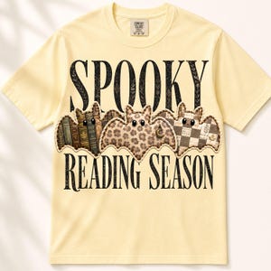 Op de afbeelding: Een lichtgeel t-shirt met korte mouwen en de zwarte tekst SPOOKY READING SEASON. Tussen de woorden staan drie vleermuisvormige afbeeldingen: één met een boekenrugpatroon, één met een luipaardprint en maanhanger, en één met een ruitjespatroon.