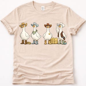 May include: A beige t-shirt with a cartoon illustration of four geese in cowboy attire. Each goose is depicted wearing a cowboy hat, boots, and other Western accessories. The design is a fun take on the cowboy theme.