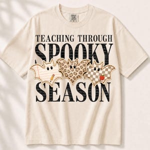 Puede incluir: Una camiseta color crema con el texto negro "TEACHING THROUGH SPOOKY SEASON" y tres gráficos en forma de murciélago. Los murciélagos tienen patrones de papel rayado con un lápiz, estampado de leopardo y un patrón de cuadros con una manzana.