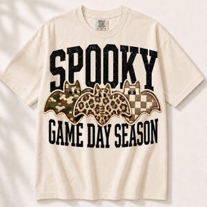 May include: A cream-colored short-sleeve t-shirt featuring the black text SPOOKY GAME DAY SEASON. Between the text are three bat illustrations: one with a green camouflage pattern, one with a brown leopard print, and one with a brown checkerboard pattern.